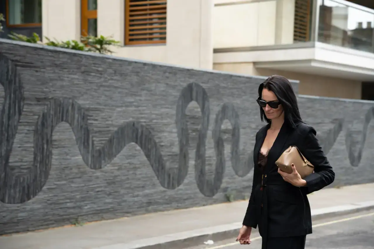 Woman in a belted black blazer holding a camel clutch, walking past a London townhouse
